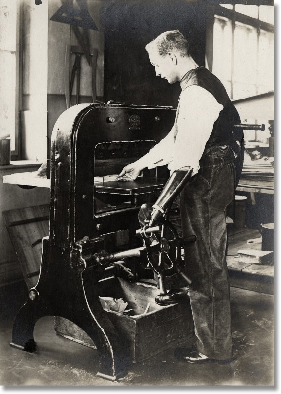 Historisches Foto eines stehenden Arbeiters mit einfacher Unterarm-Arbeitsprothese an einer Papierschneidemaschine