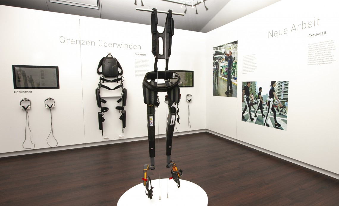 Blick in den Ausstellungsraum mit verschiedenen Exoskeletten - in der Raummitte eine frei hängende Exoskelett-Sitzhilfe mit Markenname „Noonee“ zum Selbstausprobieren