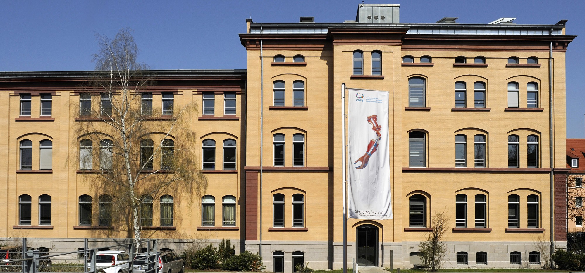 Blick auf die Fassade eines ZBFS-Gebäudes mit Eingang zur Würzburger Prothesensammlung der durch eine Fahne mit Second Hand Logo gekennzeichnet ist 