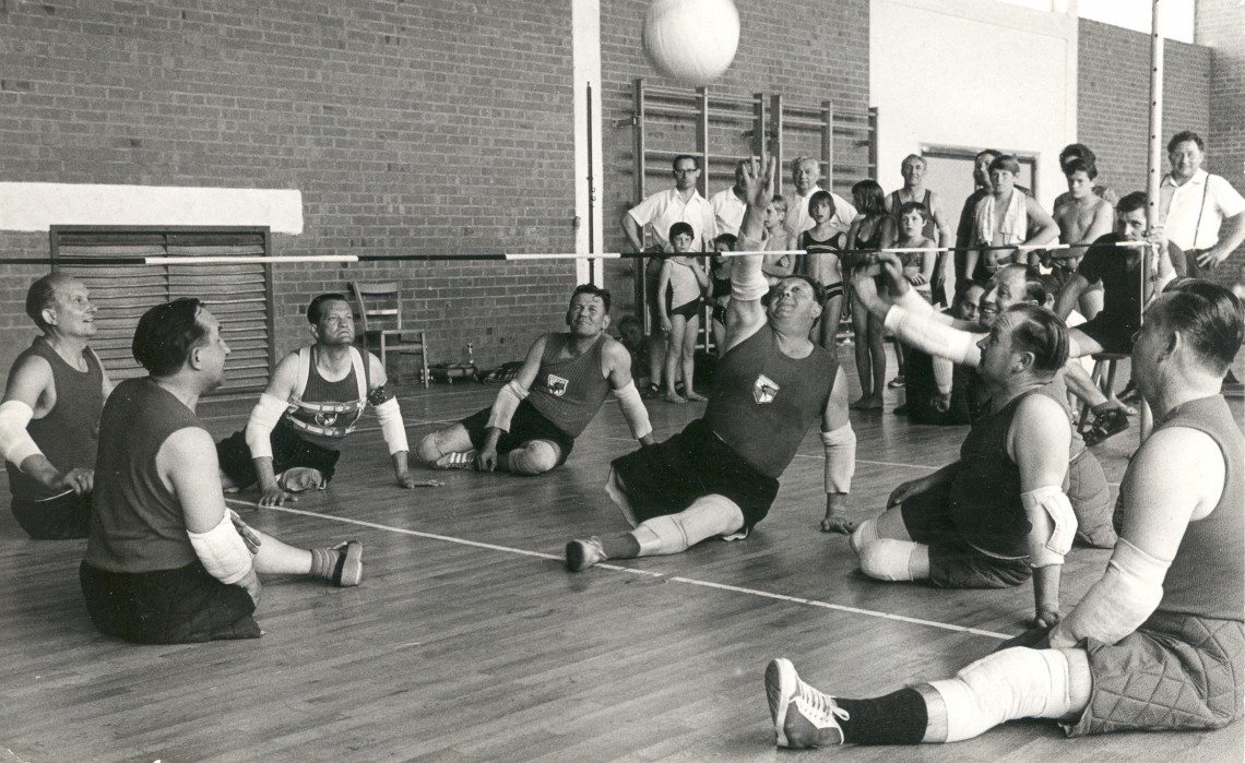 Historisches Foto aus den neunzehnhundertsechziger Jahren auf dem zwei Mannschaften beinamputierter Männer auf dem Boden sitzend gegeneinander Ball-über-die-Schnur spielen